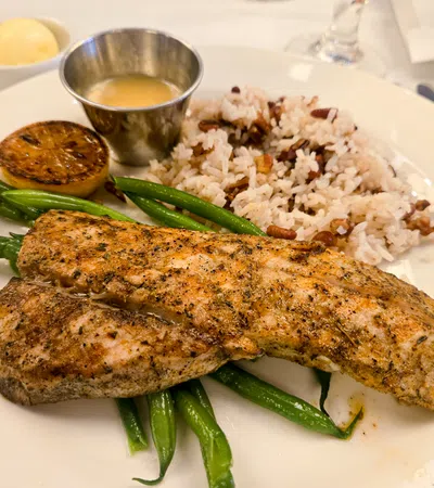 Grilled seasoned fish fillets served with green beans, rice and peas, lemon half, and sauce on a white plate.