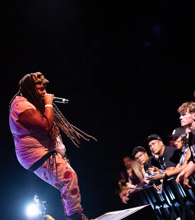 Rapper with long dreadlocks performing on stage with microphone, engaging an attentive crowd in front row