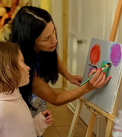 Woman teaching young girl to paint colorful balloons on canvas in indoor art class setting