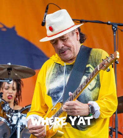 Guitarist in a white hat and yellow hoodie playing on stage with drummer in background against an orange backdrop.