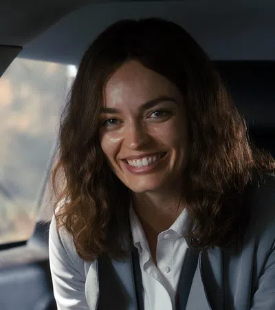 Smiling woman with curly hair wearing a blazer sitting in a car interior with natural light.