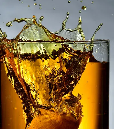 Ice cube splashing into a glass of amber-colored liquid with droplets and splashes captured mid-air.