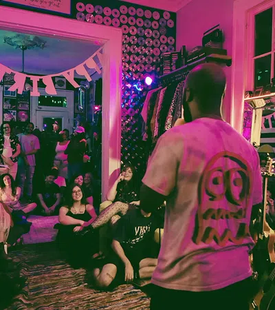 A performer entertains a seated audience in a cozy, decorated room with CDs on walls and hanging clothes.