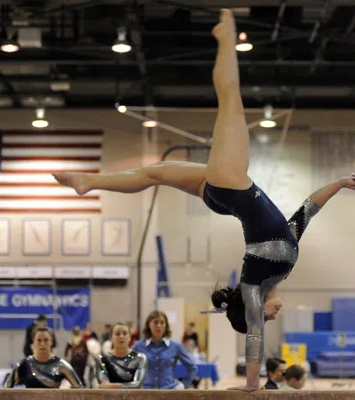 Female gymnast in blue leotard performing handstand routine at a gymnastics competition with US flag backdrop.