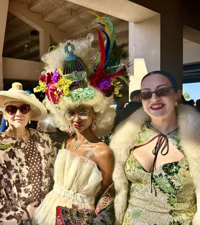 Four women in colorful, extravagant hats and elegant outfits smiling at an indoor event with natural light.