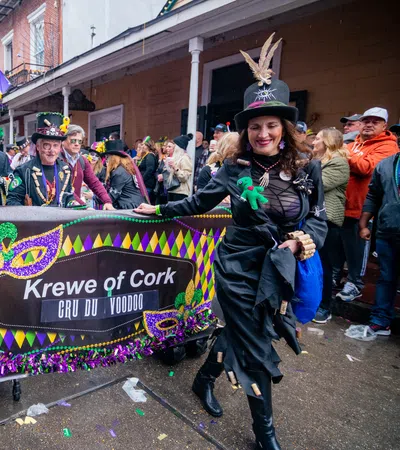 Krewe of Cork members in festive costumes holding a colorful Mardi Gras parade banner on a wet street.