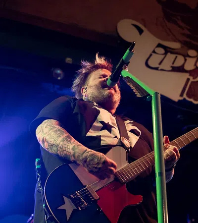 Tattooed musician passionately playing electric guitar and singing into microphone on stage with vibrant lighting.
