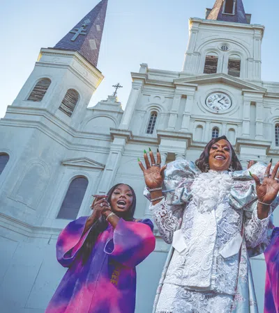 Four joyful singers in vibrant purple and white robes perform in front of a grand historic cathedral with clock tower.