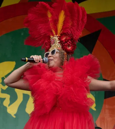 Singer in vibrant red feathered costume and oversized sunglasses performs on stage with colorful backdrop.