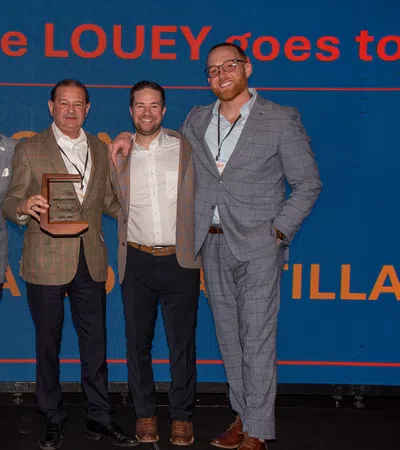 Five men in business attire pose on stage with an award plaque at a corporate event with presentation screen behind them