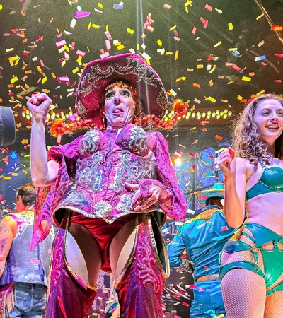 Performers in colorful costumes celebrate on stage with confetti falling and an applauding audience around them.