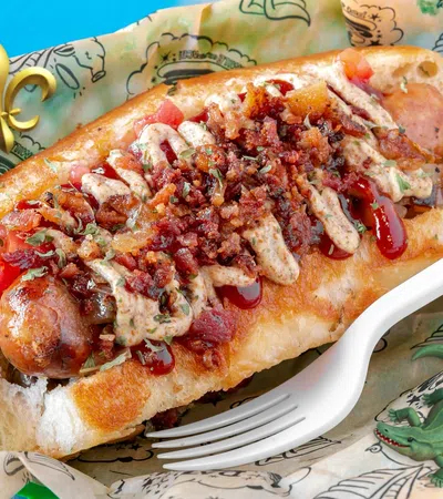 Loaded hot dog topped with crispy bacon bits, sauces, and diced tomatoes in a soft bun on patterned paper.