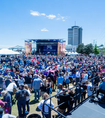 Large crowd gathered outdoors at a sunlit music festival near a river with stage and city skyline in the background