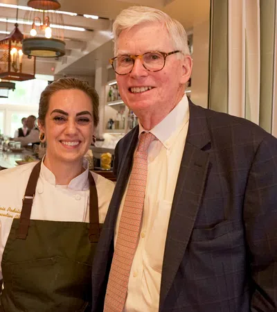 Chef and man in suit smiling inside a warmly lit restaurant with patrons at the bar in the background