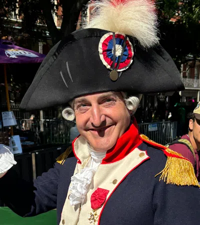 Man in historical costume smiling, dressed as a revolutionary figure with a tall hat and staff.