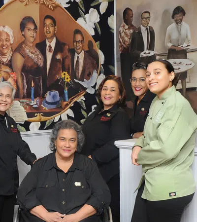 Group of diverse restaurant staff posing inside with a mural of historical figures behind them