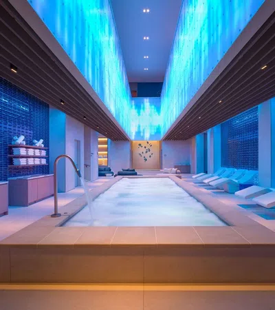 Modern indoor spa pool with blue ambient lighting, lounge chairs, and neatly stacked towels on shelves.
