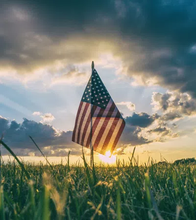 American flag standing in a grassy field with sun rays and dramatic cloudy sky at sunset