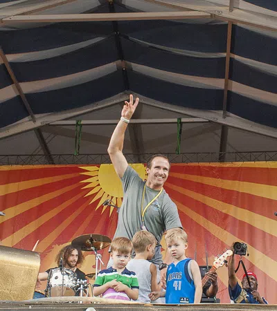 Musicians and children on stage at outdoor concert with colorful sunburst backdrop under a tent roof
