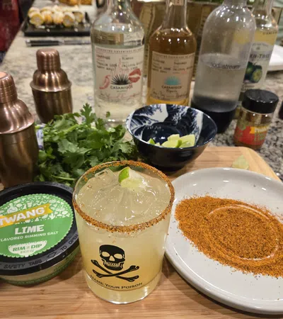 Margarita cocktail with chili rim, lime, seasoning salt, cilantro, and cocktail mixers on wooden board.