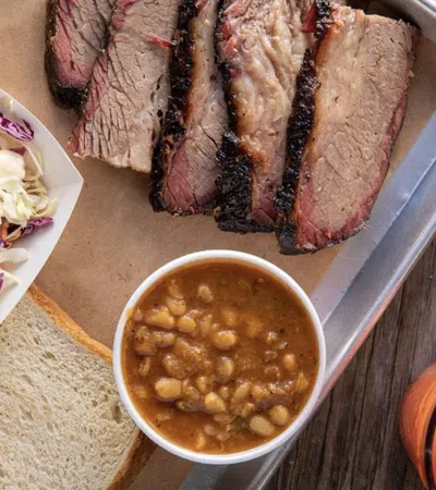 BBQ platter with sliced smoked brisket, baked beans, coleslaw, white bread, and a cocktail on a wooden table.