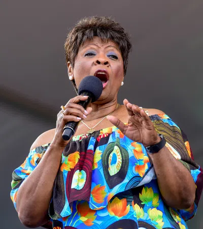 Female singer performing passionately on stage wearing a colorful off-shoulder floral dress and holding a microphone.