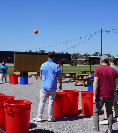 Group of men playing outdoor game tossing balls into large red bins on a sunny day with picnic tables and playground nearby