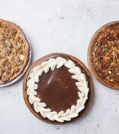 Three round pies displayed on a light surface: apple crumb, chocolate with whipped cream, and mixed nut pecan pie.