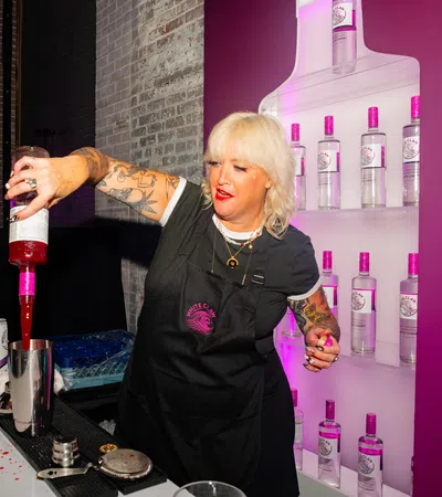 Bartender with tattoos pouring pink liquid into shaker at bar with bottles and flowers in background