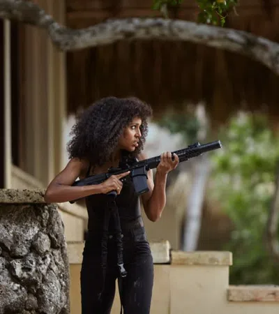 Woman with curly hair holding a rifle, standing alert beside a stone wall in an outdoor setting with trees.