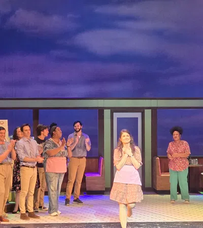 Cast of a theatrical play standing on stage with a diner set and neon pie sign in the background.