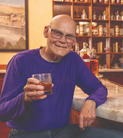 Elderly man in purple shirt sitting at bar holding glass of whiskey with shelves of liquor in background