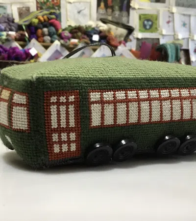 Handcrafted green knitted bus with window patterns and button wheels displayed on a white table in a craft store.
