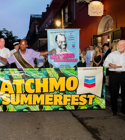 People holding a colorful Satchmo SummerFest banner during an evening street event with buildings and onlookers.