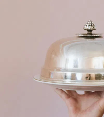 Person holding a silver cloche food cover on a tray against a soft pink background