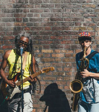 Two street musicians perform against a brick wall, one playing electric guitar and singing, the other playing saxophone.