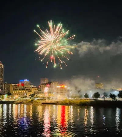 Colorful fireworks burst above a city skyline at night reflecting on calm river water below.