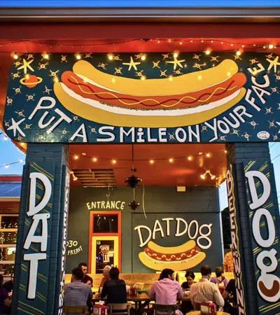 Front entrance of Dat Dog restaurant with colorful hot dog sign and dining patrons at dusk under string lights