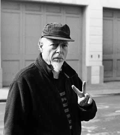 Black and white photo of a man with a goatee wearing a cap and jacket showing a peace sign outdoors on street.