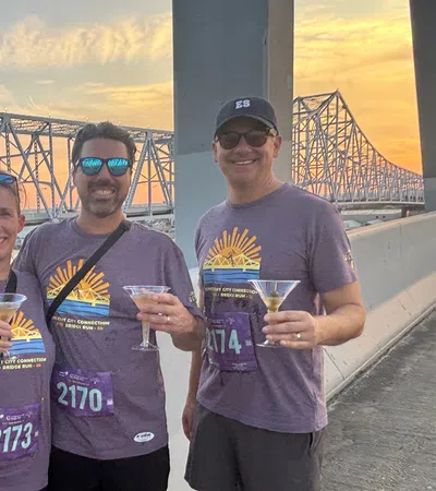 Four runners wearing matching purple shirts hold drinks by a bridge at sunset celebrating their run.