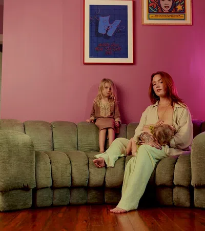 Mother breastfeeding baby on green couch while young girl sits beside them in pink room with framed art.