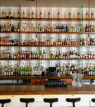 Wide bar with rows of liquor bottles on shelves and empty black bar stools lined up at wooden counter.