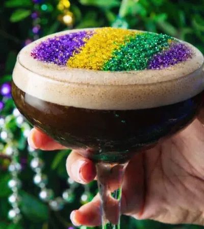 Hand holding a cocktail glass with dark drink topped with purple, yellow, and green glitter foam for Mardi Gras celebration.