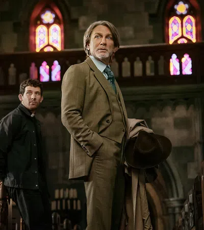Two men, one in a suit holding a hat, standing inside a church with stained glass windows and wooden pews.