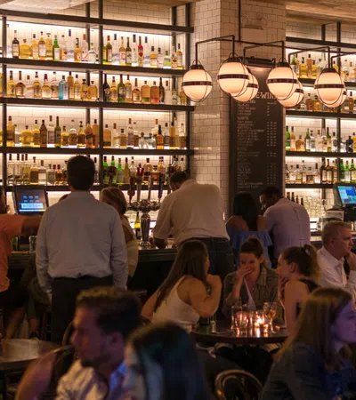 Busy bar scene with patrons sitting and standing, illuminated bottles on shelves, and hanging globe lights.