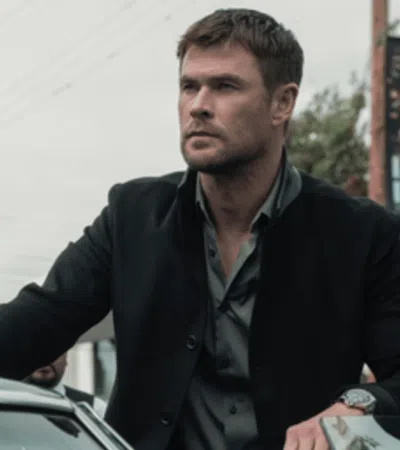 Man in black jacket leaning on a vintage car in an urban street setting with utility poles in the background.