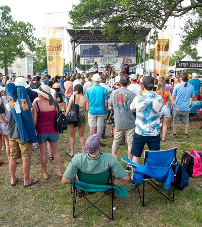 Crowd of people enjoying outdoor music festival with stage, banners, and attendees in casual summer attire.