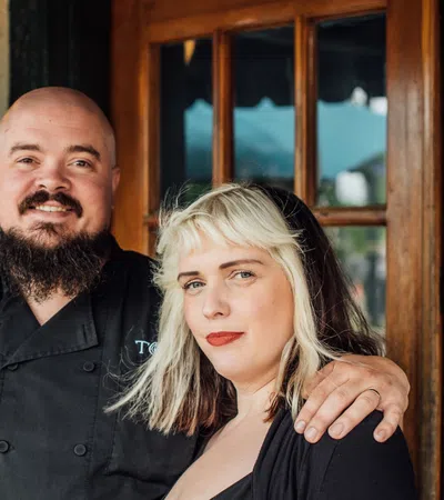 Smiling chef with beard in black uniform embraces woman with blonde hair outside restaurant entrance.