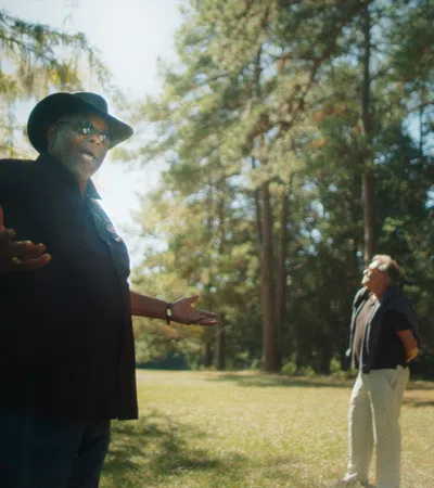 Man in black hat and shirt speaking to three men standing in a sunlit forest clearing.