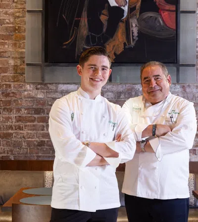 Two chefs in white uniforms standing in a rustic restaurant with brick walls and a chef portrait painting.